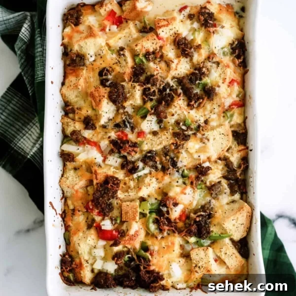 Baked casserole with bread cubes, bell peppers, onions, melted cheese, and browned sausage in a white rectangular dish on a green checkered cloth.