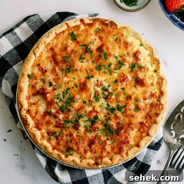 A golden-brown quiche garnished with chopped herbs sits on a checkered cloth. A pie server is beside it, with small bowls of chopped herbs and strawberries nearby.