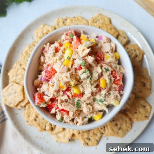 white bowl surrounded by crackers with tuna corn salad topped with herbs, red bell pepper, and onion