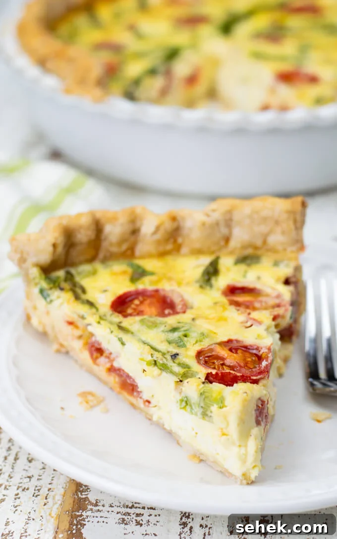 Asparagus and Cherry Tomato Quiche Recipe for Spring Brunch 8 A piece of asparagus quiche with tomatoes sits on a white plate with a fork next to it. The pie pan with quiche sits blurred in the background.