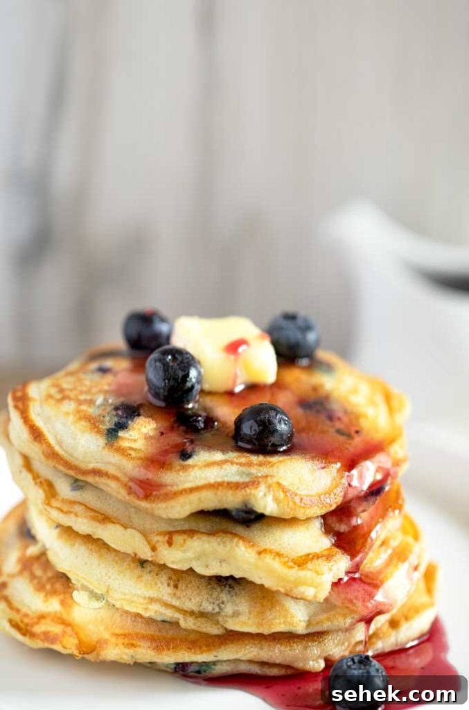 Fluffy Blueberry Pancakes (Made with Frozen Berries) 2 A stack of blueberry pancakes with butter and blueberry syrup on a white plate.