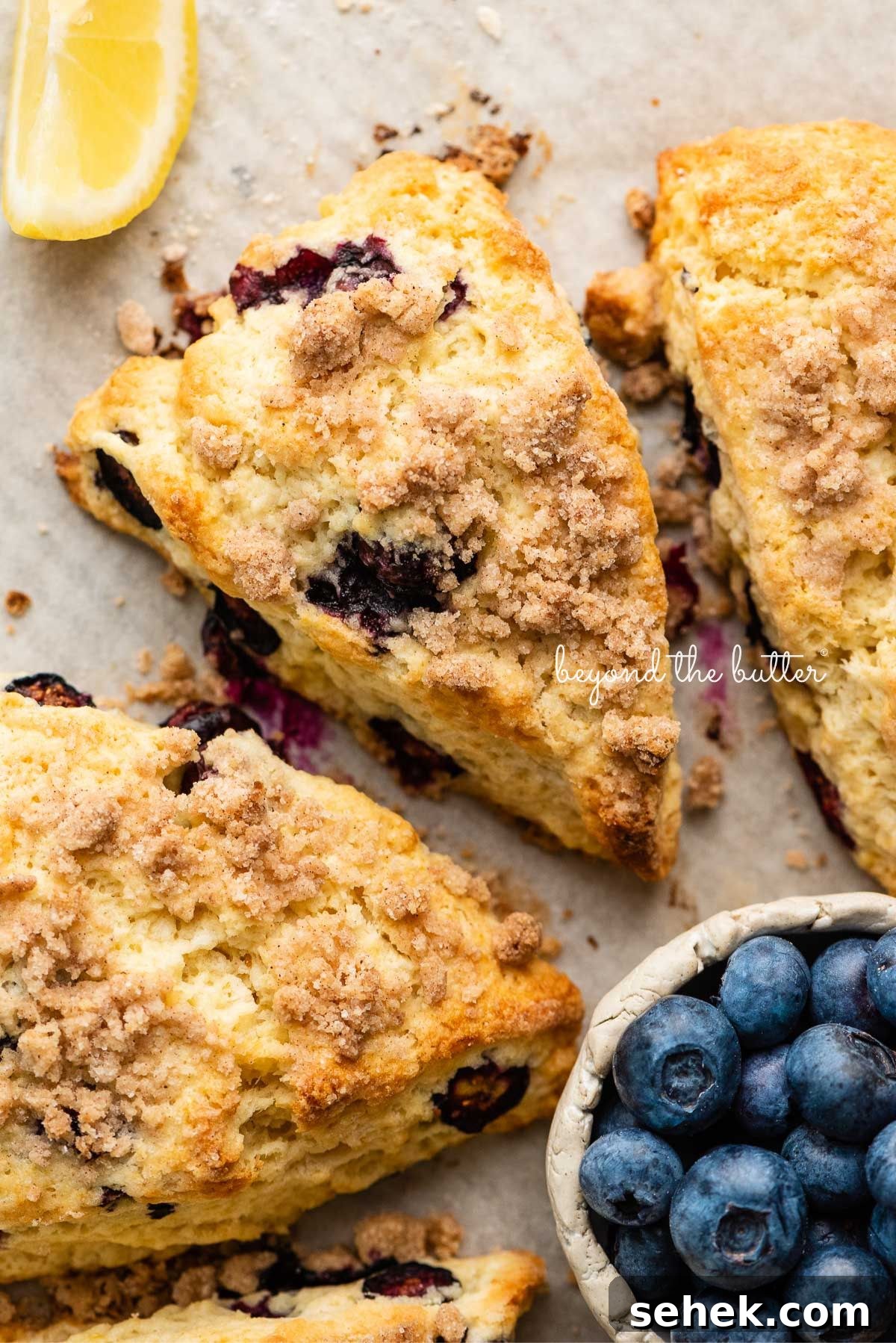 Lemon blueberry streusel scones on a parchment lined baking sheet placed next to a lemon wedge and small ceramic bowl of blueberries.