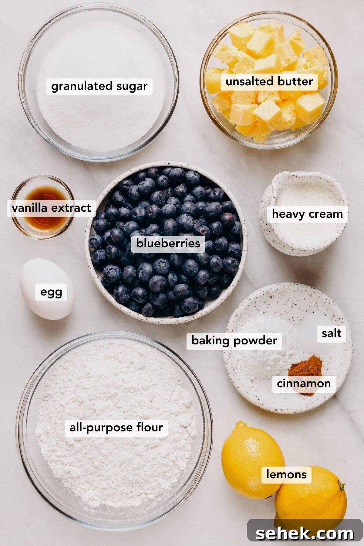 Ingredients in various size bowls labeled as granulated sugar, unsalted butter, vanilla extract, blueberries, heavy cream, egg, baking powder, salt, cinnamon, all-purpose flour, and lemons on a white marbled background.