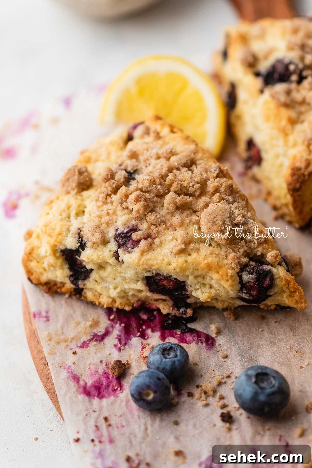 Lemon blueberry scones with streusel topping placed on a parchment paper lined round wooden serving plate with lemon wedges and blueberries placed around it.