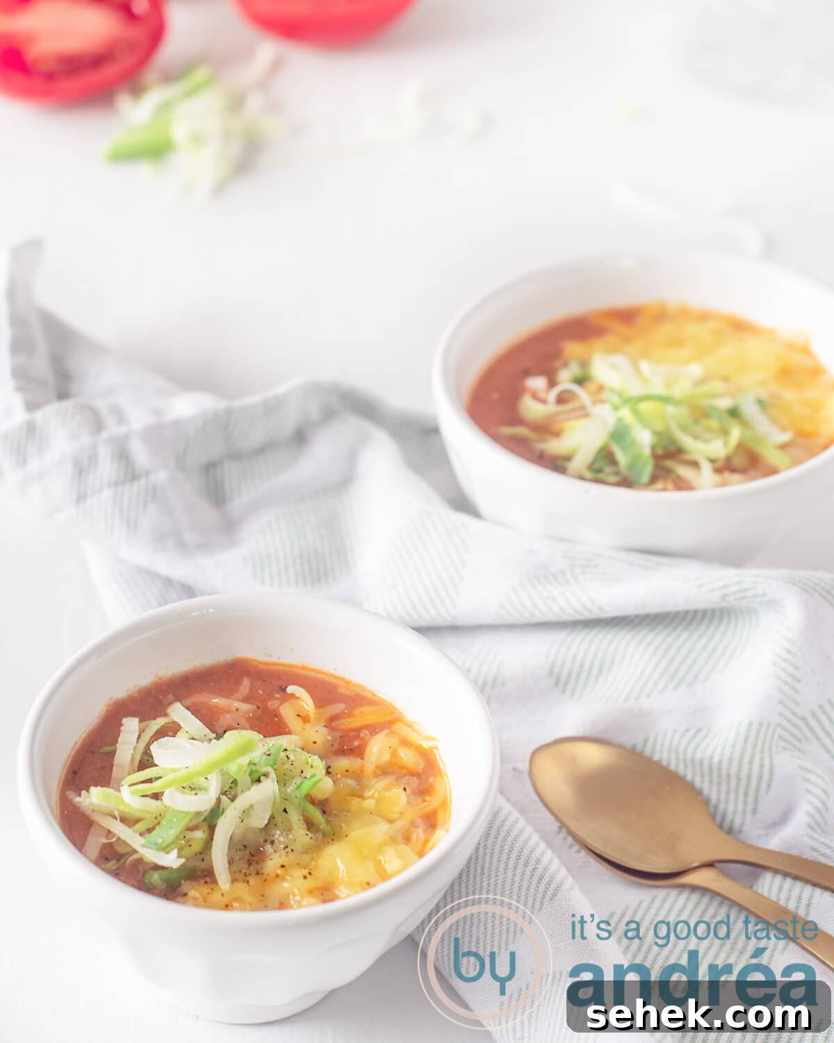 Slow-Cooker Creamy Tomato Soup Recipe for Cozy Weeknights 2 Two bowls filled with tomato soup with a cheese and leek topping. Two gold spoons on the right. A white green tea towel lies between the bowls.