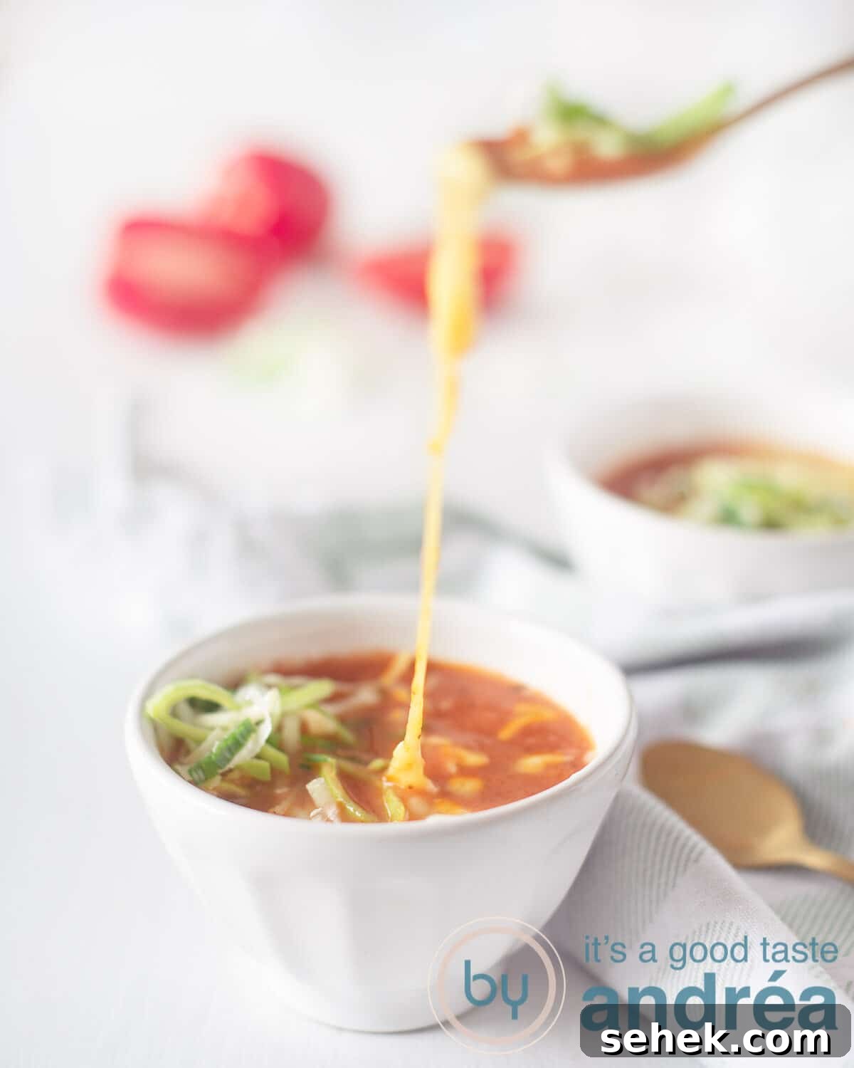 Slow-Cooker Creamy Tomato Soup Recipe for Cozy Weeknights 3 Two bowls of tomato soup (one front and center) with leeks and cheese. A spoon takes a bite and melted cheese hangs between the soup and the spoon. A green and white tea towel lies between the bowls with two golden spoons on top. diced tomatoes in the background