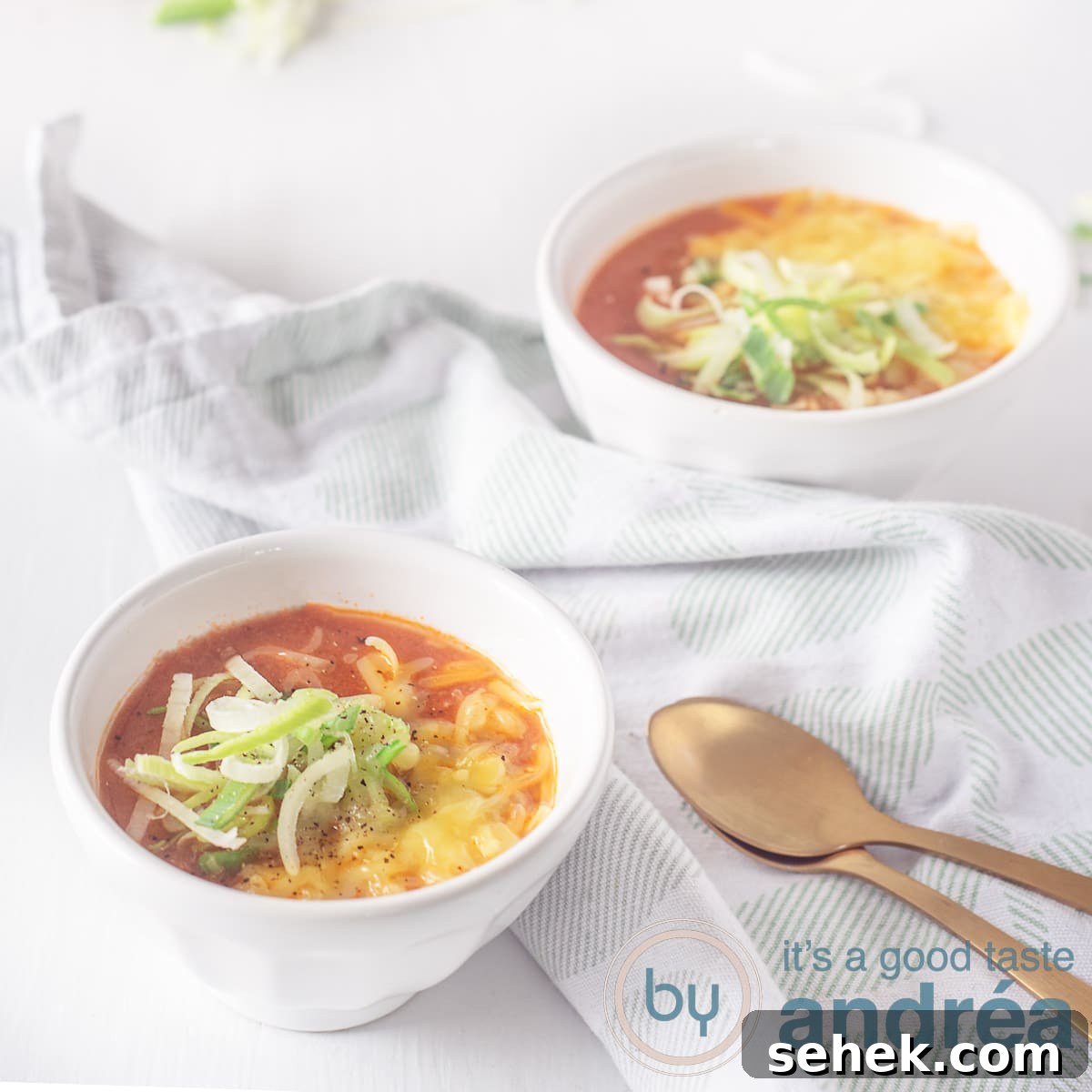 Slow-Cooker Creamy Tomato Soup Recipe for Cozy Weeknights 4 A square photo of a white background with two white bowls filled with tomato soup. With a topping of leek and cheese. Two golden spoons at the bottom left