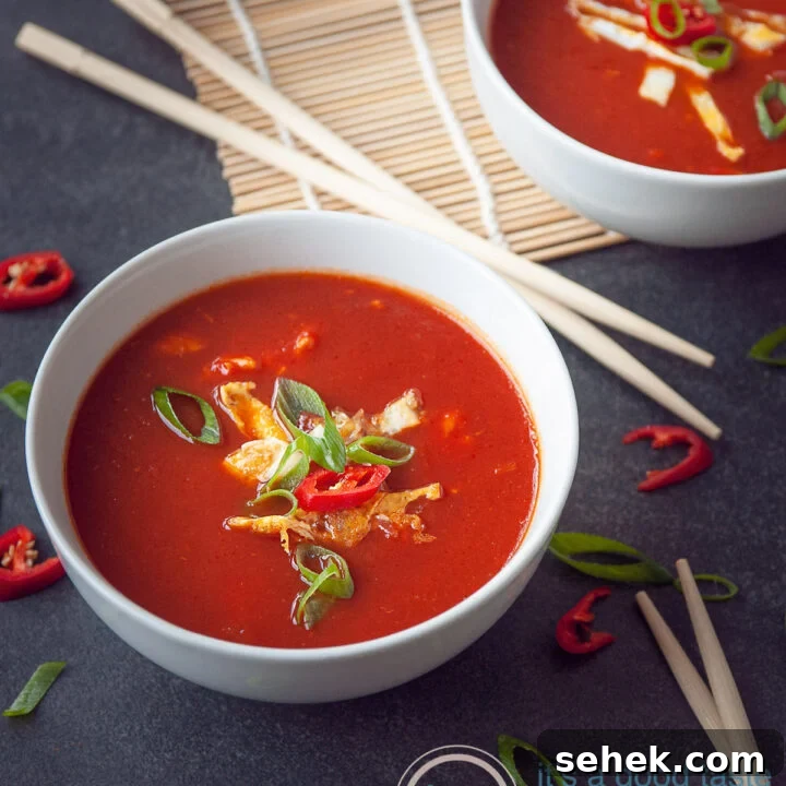 A grey background with two bowls filled with Asian tomatosoup with egg, spring onions and pepper. Some chopsticks and veggies scattered around