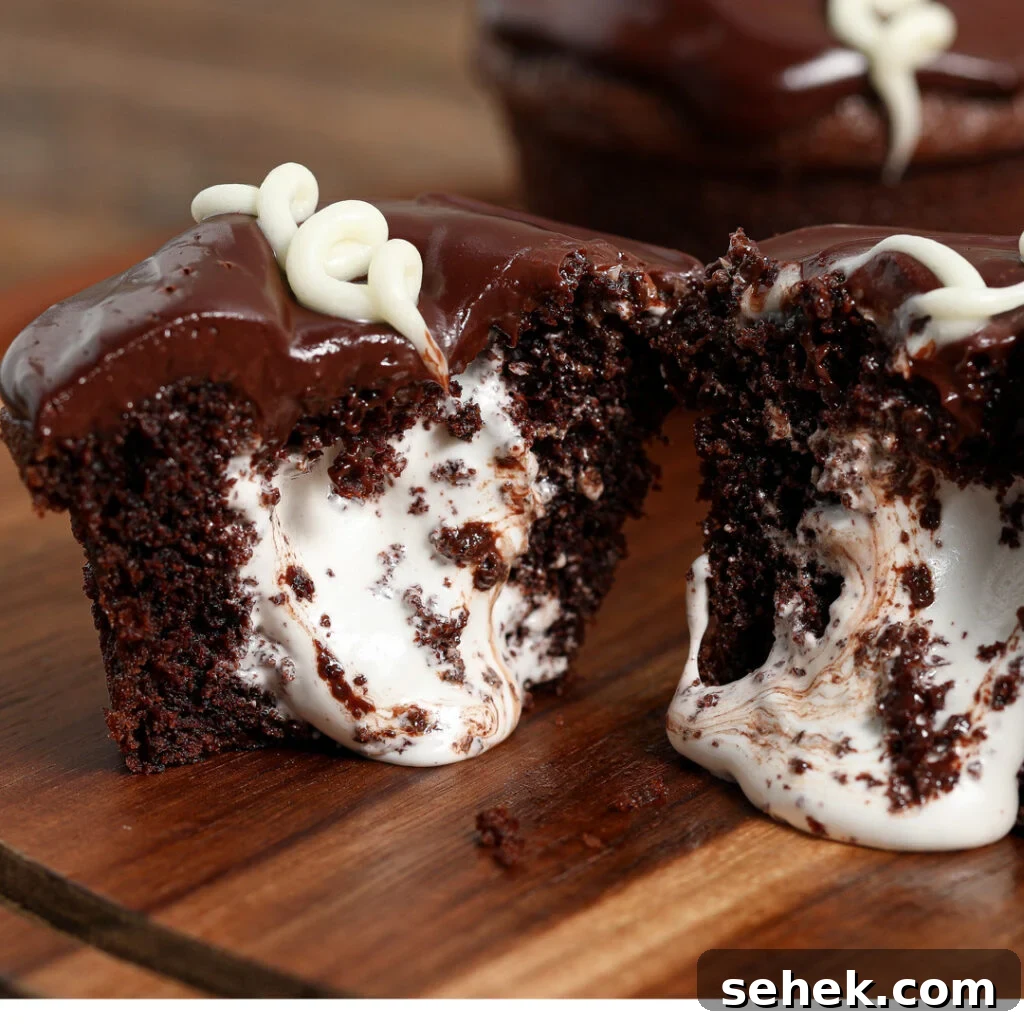 Copycat Little Debbie Oatmeal Creme Cupcakes Recipe 2 A homemade Little Debbie Cupcake cut in half reveals a creamy white filling inside. Topped with glossy chocolate icing and a swirl of white icing, the cupcake sits on a wooden surface.