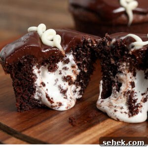 Copycat Little Debbie Oatmeal Creme Cupcakes Recipe 4 A homemade Little Debbie Cupcake cut in half reveals a creamy white filling inside. Topped with glossy chocolate icing and a swirl of white icing, the cupcake sits on a wooden surface.