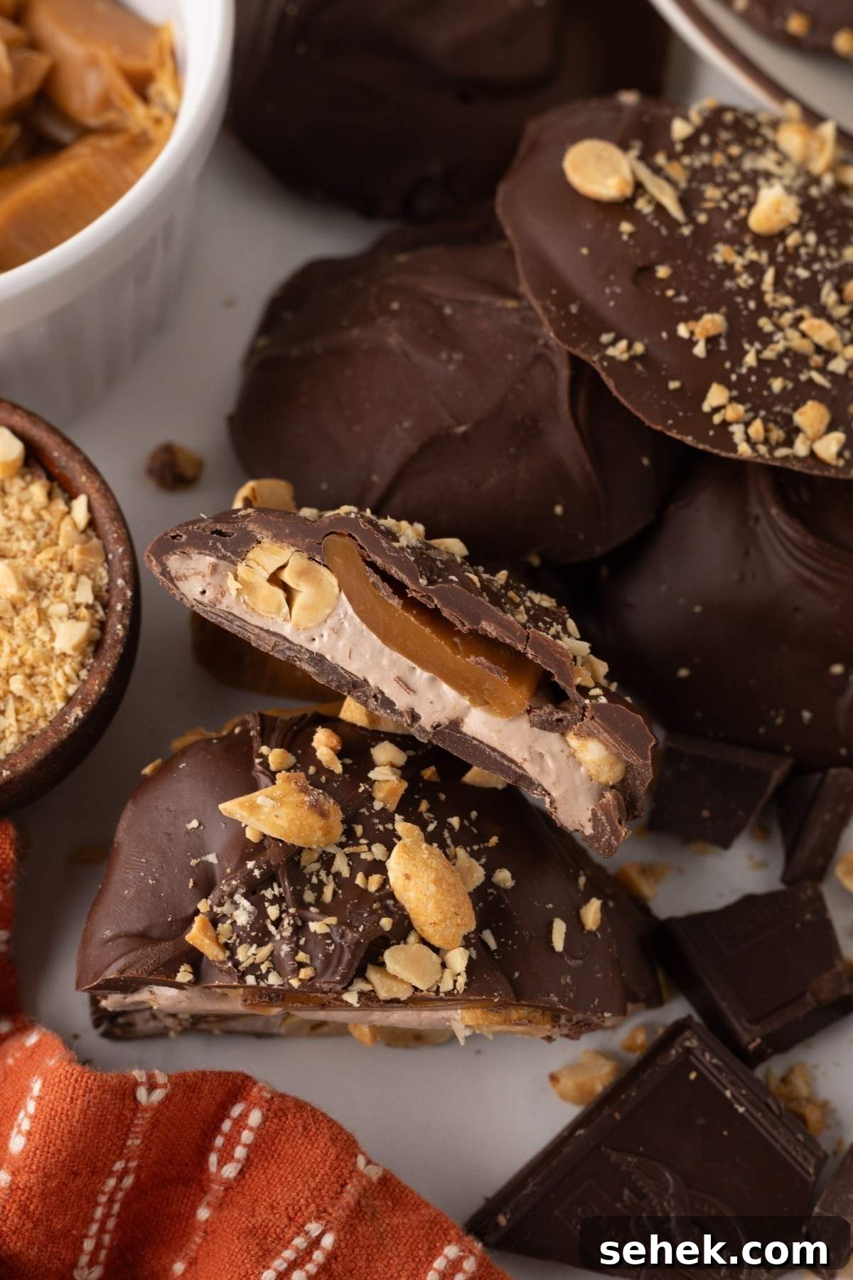 Homemade Snickers bites with a layer of caramel and peanuts, adorned with chocolate, are arranged artfully with some broken pieces and a small bowl of crushed peanuts nearby. An orange-striped cloth is partially visible, adding a touch of warmth.