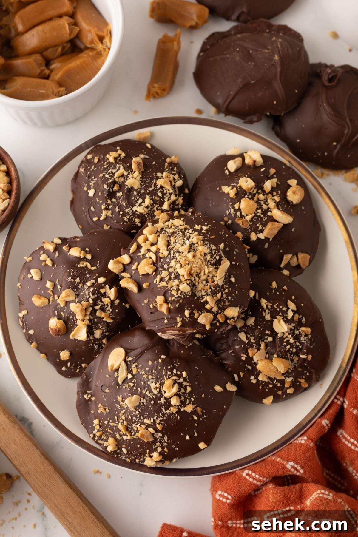 A plate of chocolate-covered cookies topped with chopped nuts, reminiscent of homemade Snickers bites. Additional cookies and caramel pieces are scattered around on a marble surface.