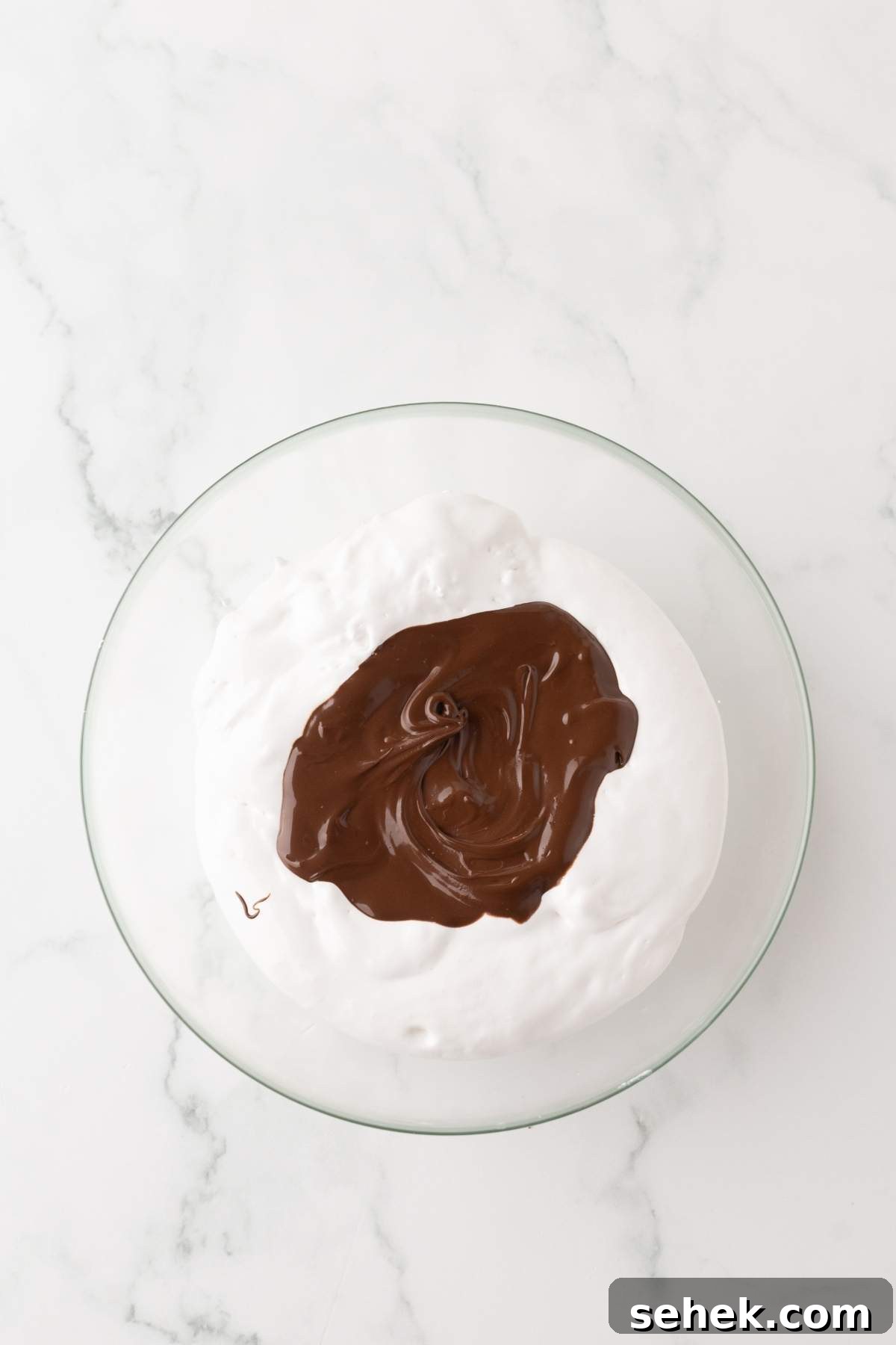 A glass bowl filled with white whipped cream, topped with a dollop of melted chocolate and garnished with crunchy homemade Snickers bites, sits elegantly on a marble surface.