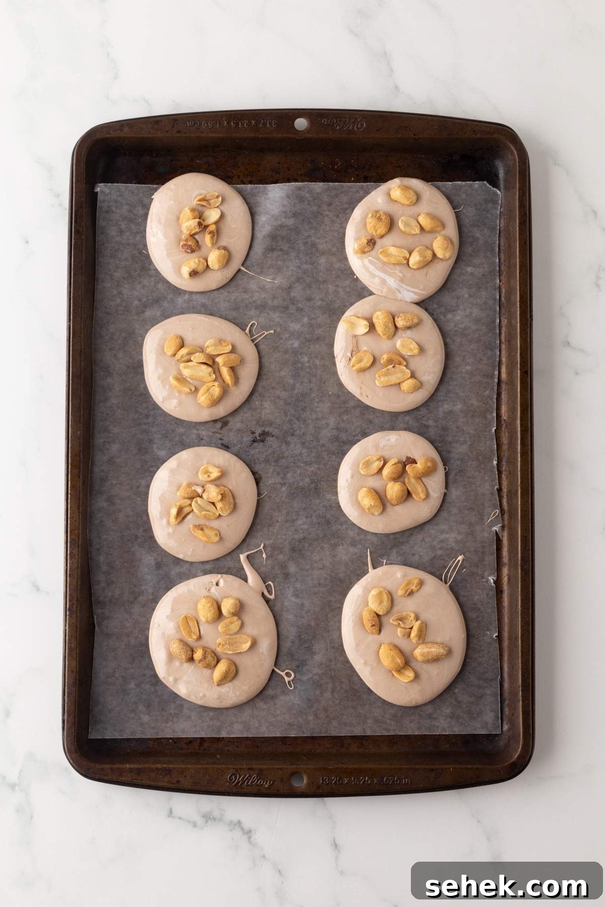 A baking tray with eight round dough pieces topped with peanuts on parchment paper, resembling delightful homemade Snickers bites.