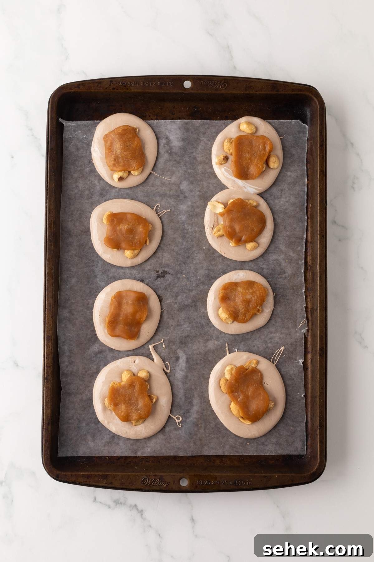 Unbaked cookies with caramel and nuts, reminiscent of homemade Snickers bites, are arranged on a parchment-lined baking sheet.