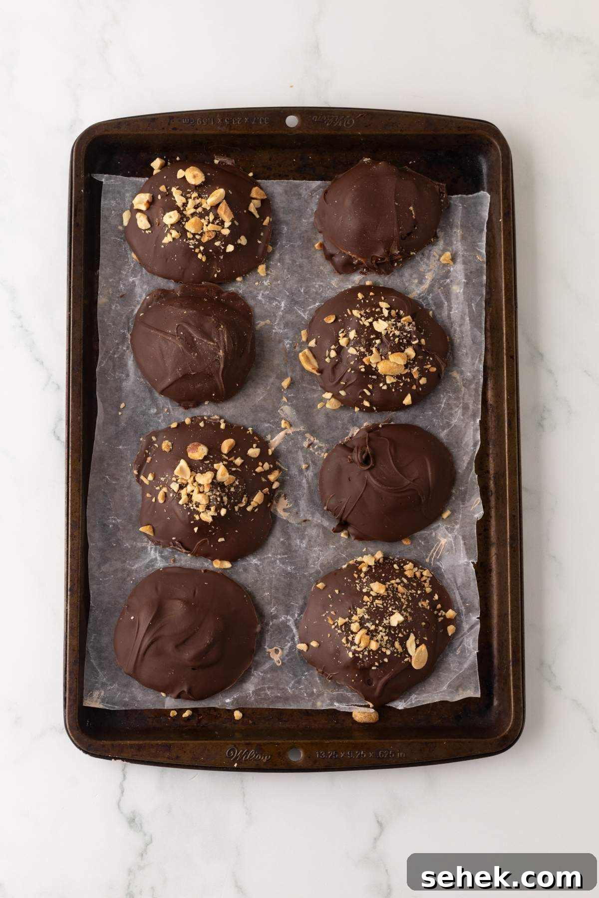 A baking tray with eight homemade Snickers bites, featuring chocolate-covered cookies topped with crushed nuts, all perfectly arranged on parchment paper.