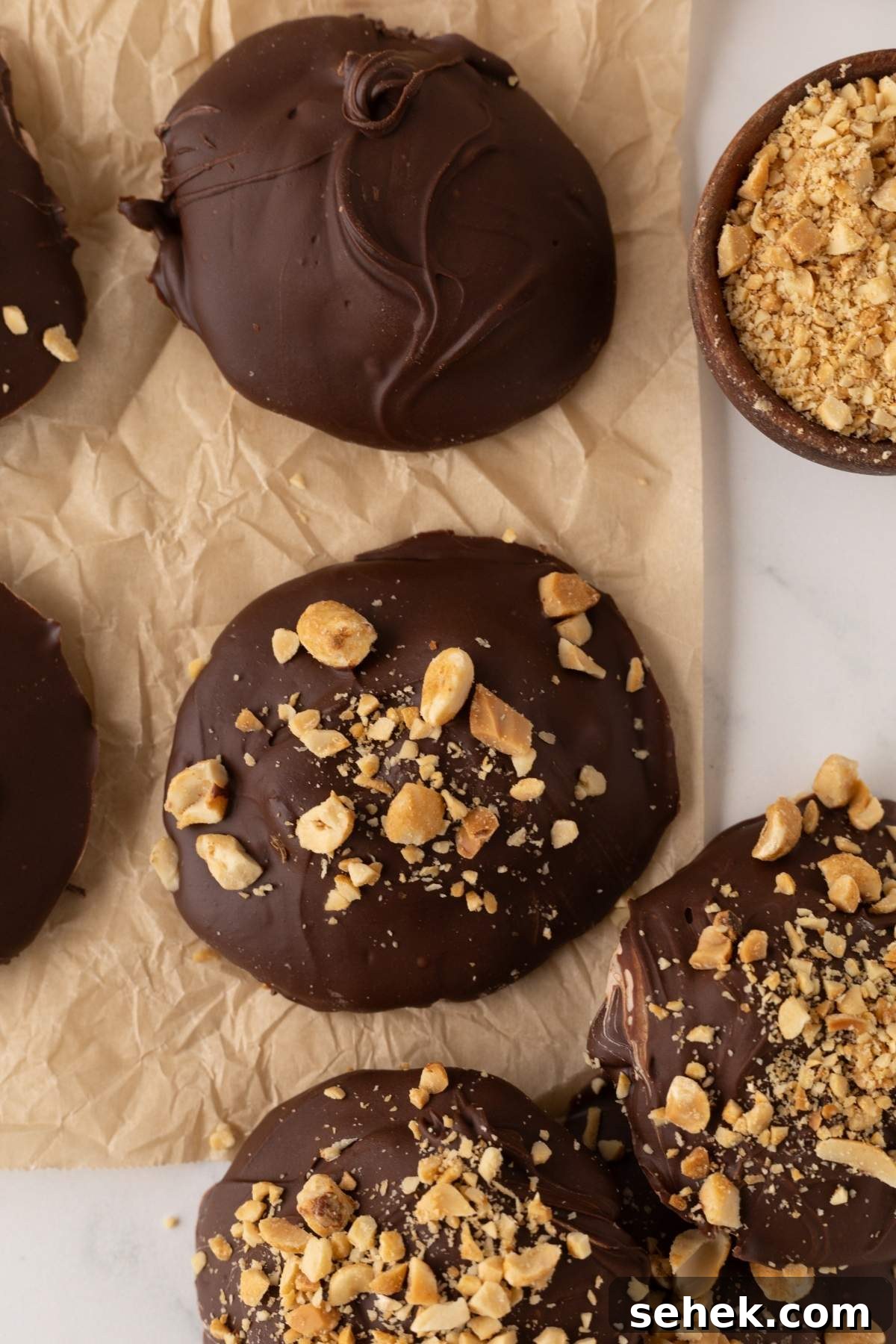 Homemade Snickers bites, these chocolate-covered cookies are topped with crushed peanuts on parchment paper, accompanied by a small bowl of extra crushed peanuts.