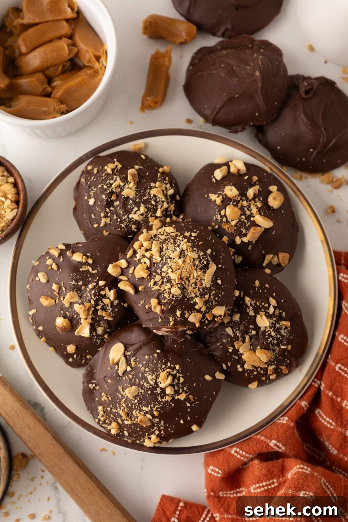 A plate of homemade Snickers bites topped with chopped nuts, surrounded by caramel pieces, nuts, and a red cloth.