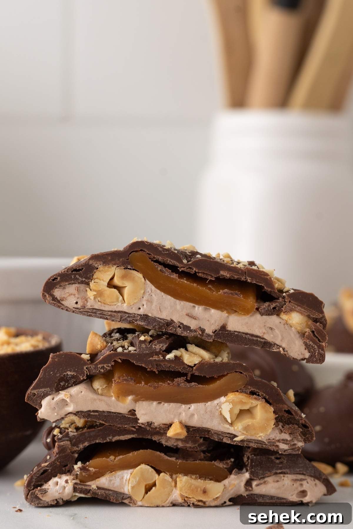 Close-up of stacked homemade Snickers bites brimming with caramel and peanuts. A blurred container with kitchen utensils sits in the background, adding a cozy kitchen vibe.