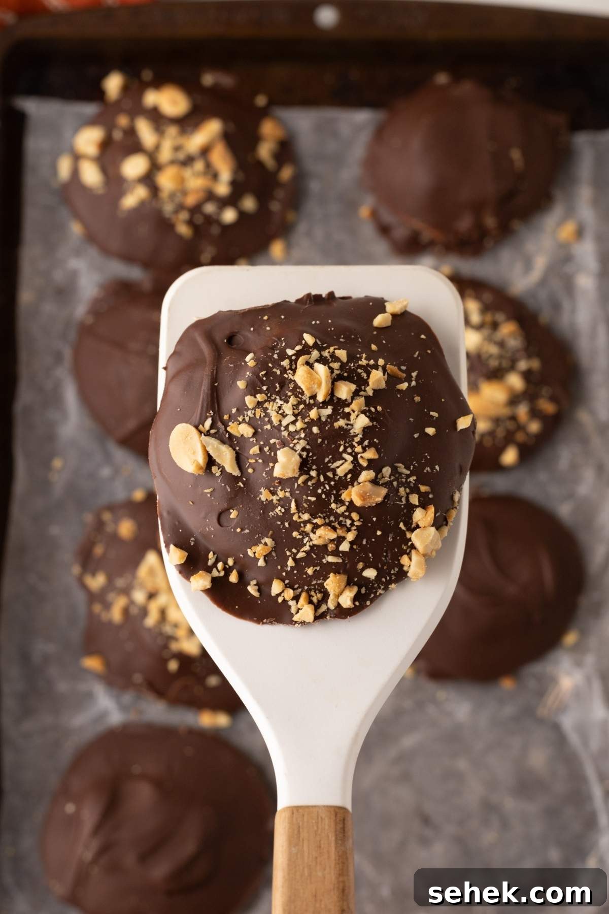 A spatula holds a homemade Snickers bite, its chocolate-coated surface adorned with chopped nuts, while more of these delightful bites rest on a baking sheet in the background.