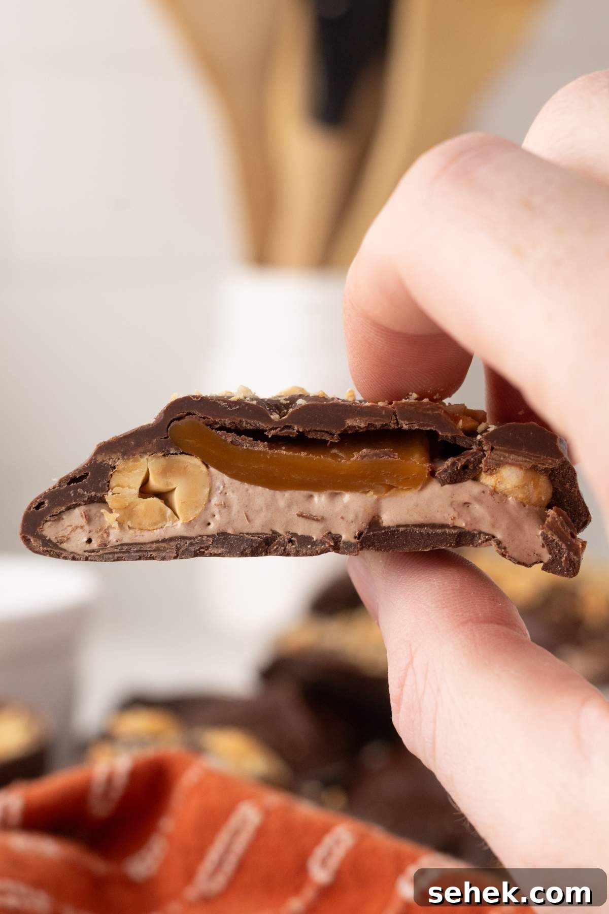 Close-up of a hand holding a homemade Snickers bite, showcasing rich chocolate encasing caramel, nuts, and nougat layers, with a blurred kitchen setting in the background.