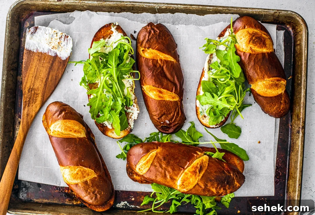 Brie and Roast Beef Sandwiches with Savory Au Jus 4 Sheet pan holding toasted pretzel buns topped with arugula with a spatula smeared with creamy horseradish sauce.