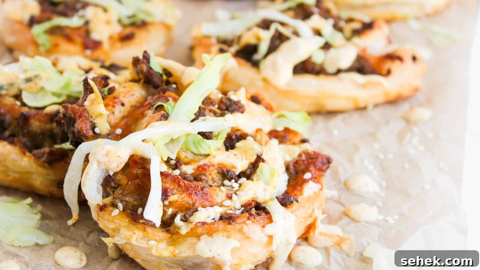 9 Pinwheel Recipes Guests Always Request Again 3 Close-up of puff pastry rounds topped with ground meat, shredded lettuce, sesame seeds, and a creamy sauce, arranged on parchment paper.