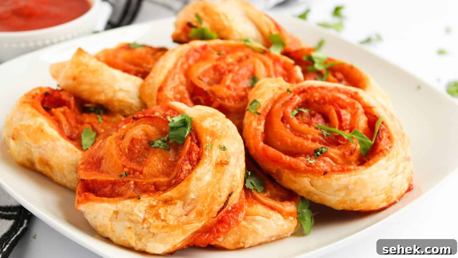 9 Pinwheel Recipes Guests Always Request Again 5 A plate of golden-brown pinwheel pastries filled with tomato sauce and garnished with fresh herbs. A small bowl of sauce is in the background.
