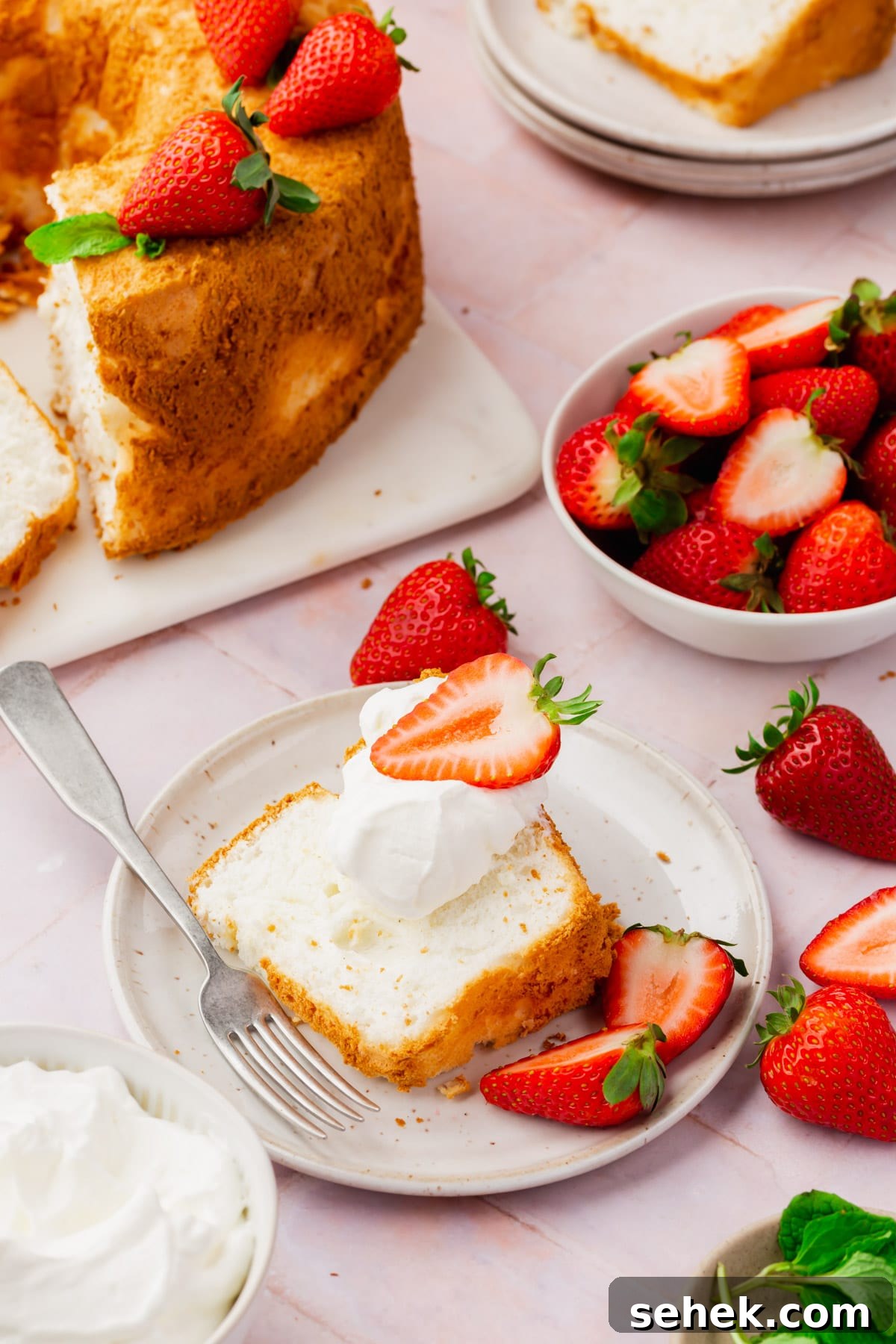 Light and Fluffy Gluten-Free Angel Food Cake Recipe 5 A slice of angel food cake on a plate topped with whipped cream and a strawberry with a full angel food cake and a bowl of strawberries in the background.