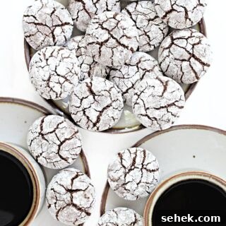 Plate of chocolate crinkle cookies covered in powdered sugar with cracks showing chocolate cookie inside.