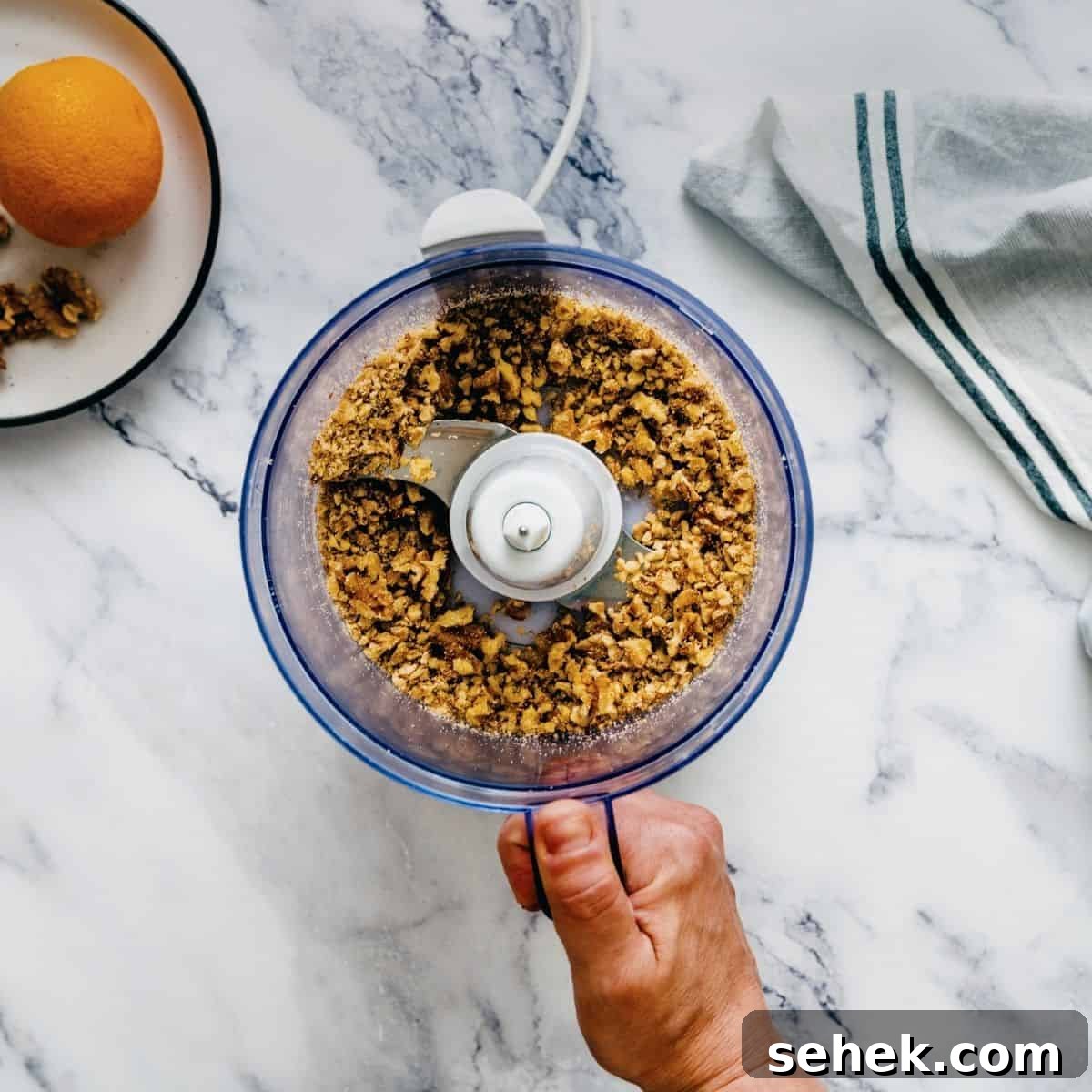 Melomakarona Greek Christmas Honey Cookies Recipe 5 a hand holding a bowl of a food processor with crushed walnuts.
