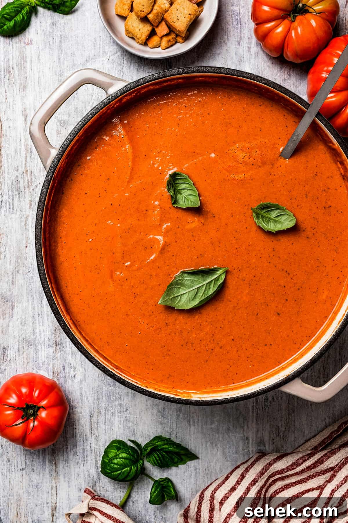 This creamy tomato bisque is delicious and so easy to make with canned tomatoes, heavy cream, and Parmesan cheese. A perfect soup for lunch or dinner. overhead shot of a soup pot with tomato bisque, topped with three basil leaves, and tomatoes and croutons placed around the pot.