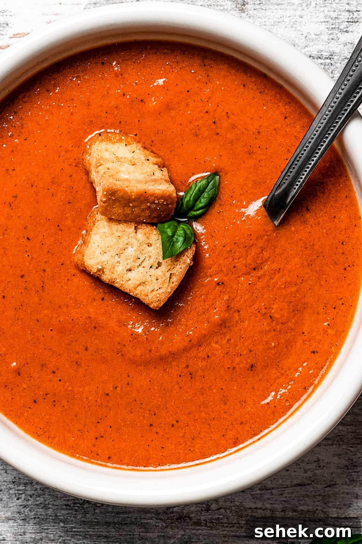 This creamy tomato bisque is delicious and so easy to make with canned tomatoes, heavy cream, and Parmesan cheese. A perfect soup for lunch or dinner. close up shot of a bowl of creamy tomato bisque garnished with croutons and basil leaves, a spoon stirring through it.