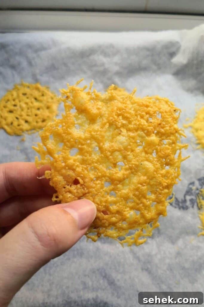 Hand holding up a parmesan crisp with more parmesan crisps on an oven tray in the background.