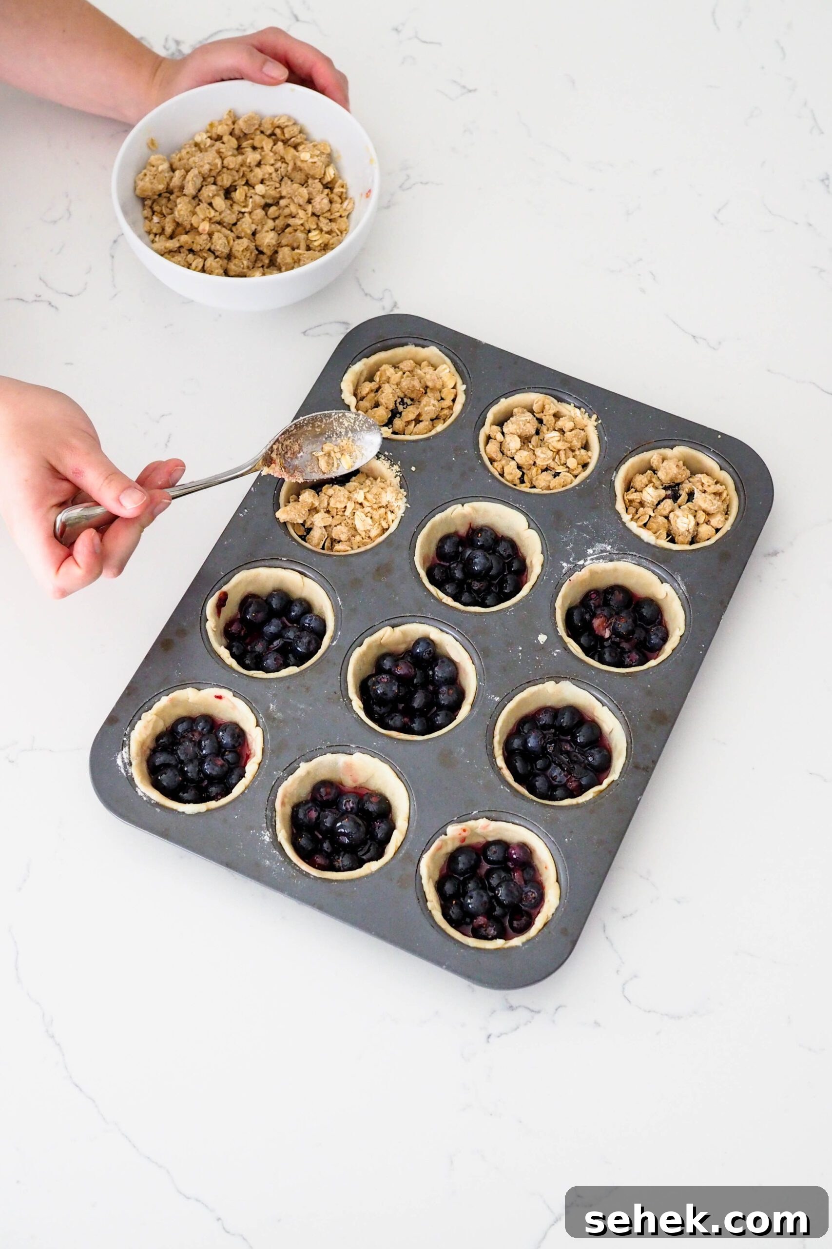 Mini Blueberry Hand Pies: Bite-Sized Fruit Desserts for Any Occasion 9 A hand spoons crumble onto mini blueberry pie shells.