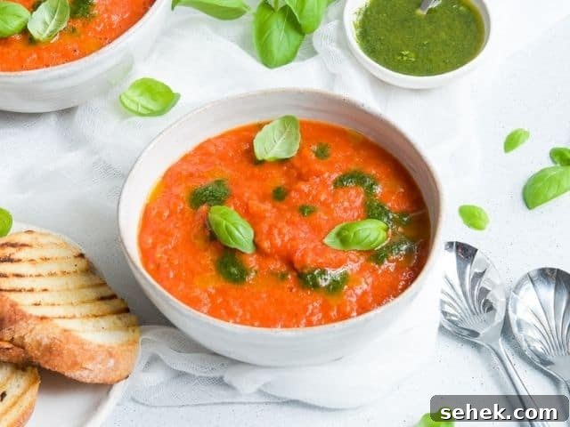 bowl of soup, garnished with basil leaves, surrounded by some toasted bread, spoons and more basil leaves, another bowl of soup and dish of basil oil in the background.