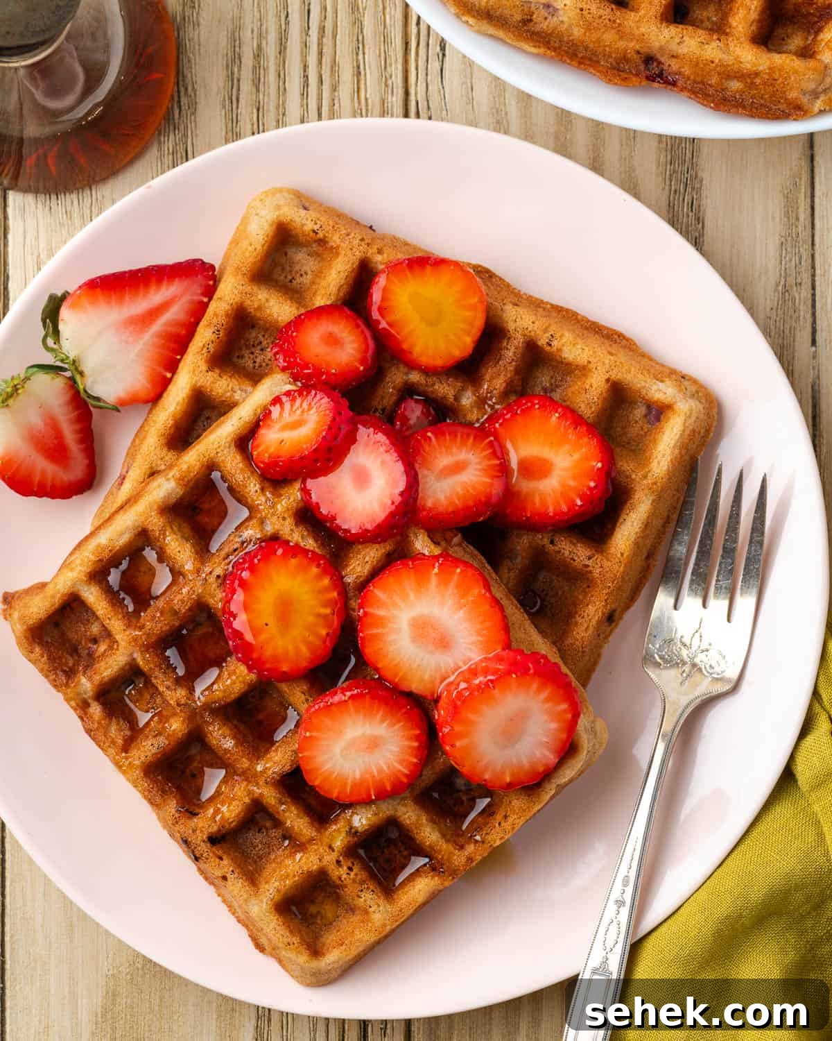 how to make strawberry waffles Two gluten free strawberry waffles served on a plate topped with fresh strawberry slices and syrup.