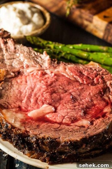 a slice of prime rib on a white plate with mashed potatoes and grilled asparagus, next to a small dish of horseradish sauce. the rest of the standing rib roast is in the background
