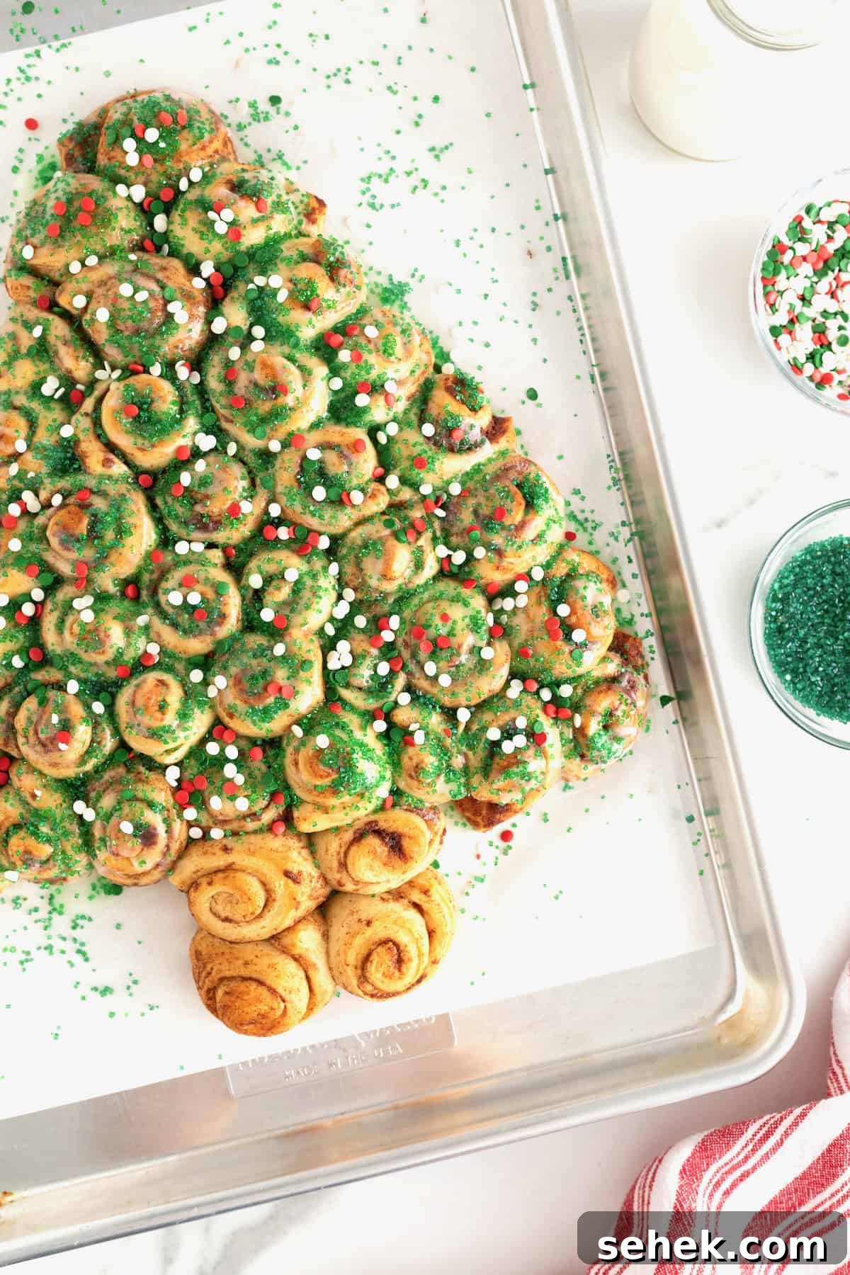 Cinnamon Roll Christmas Tree Pull-Apart Recipe 11 A cinnamon roll Christmas tree sprinkled with green sugar crystals and red and green holiday sprinkles on a parchment lined baking sheet.