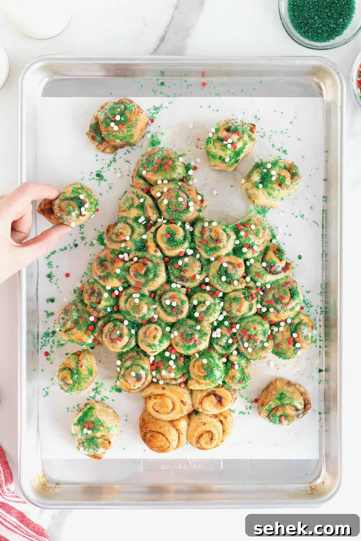 Cinnamon Roll Christmas Tree Pull-Apart Recipe 3 A hand taking a piece of a cinnamon roll pull-apart shaped like a Christmas tree topped with holiday sprinkles.