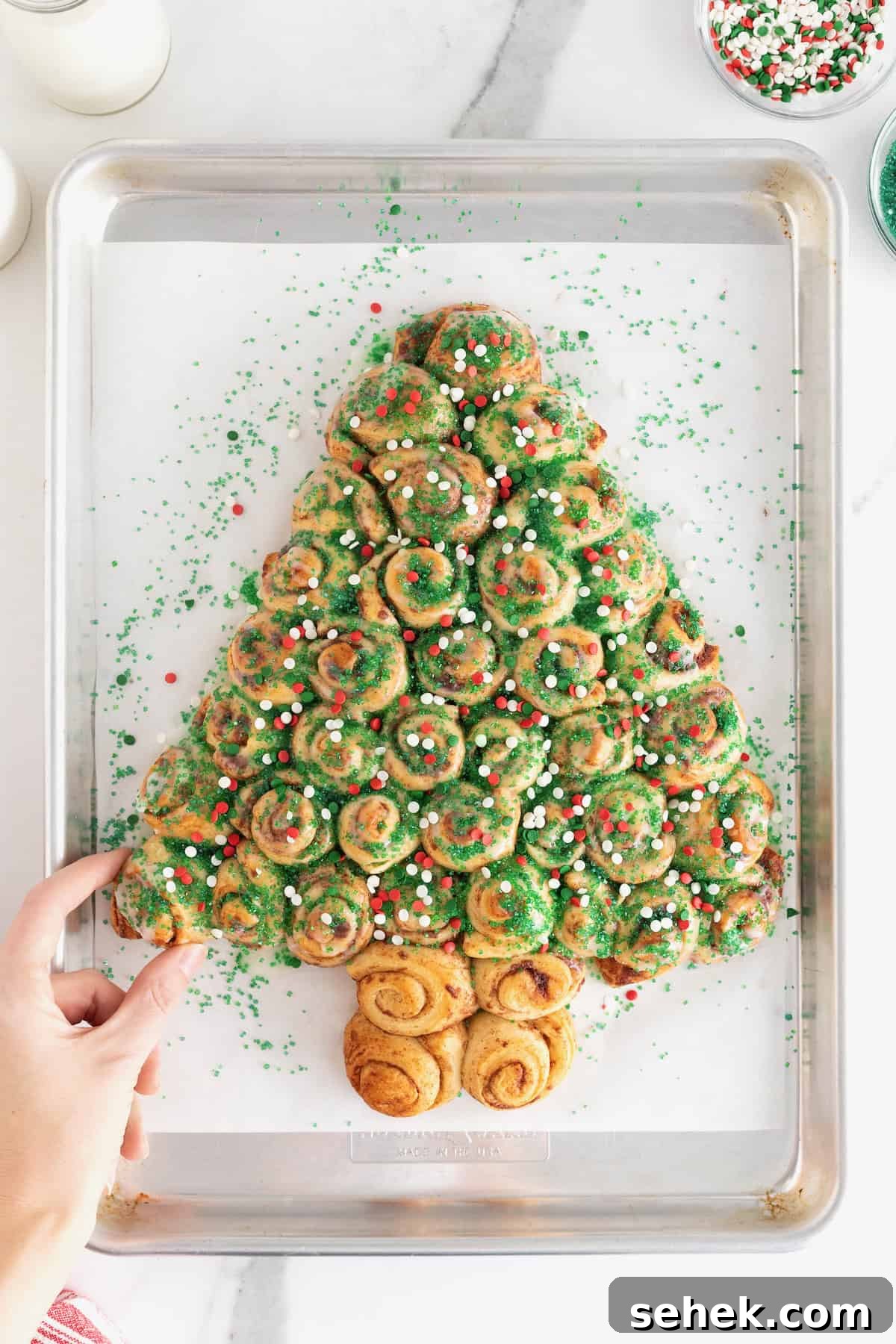 Cinnamon Roll Christmas Tree Pull-Apart Recipe 4 A hand taking a piece of a cinnamon roll pull-apart shaped like a Christmas tree topped with holiday sprinkles.