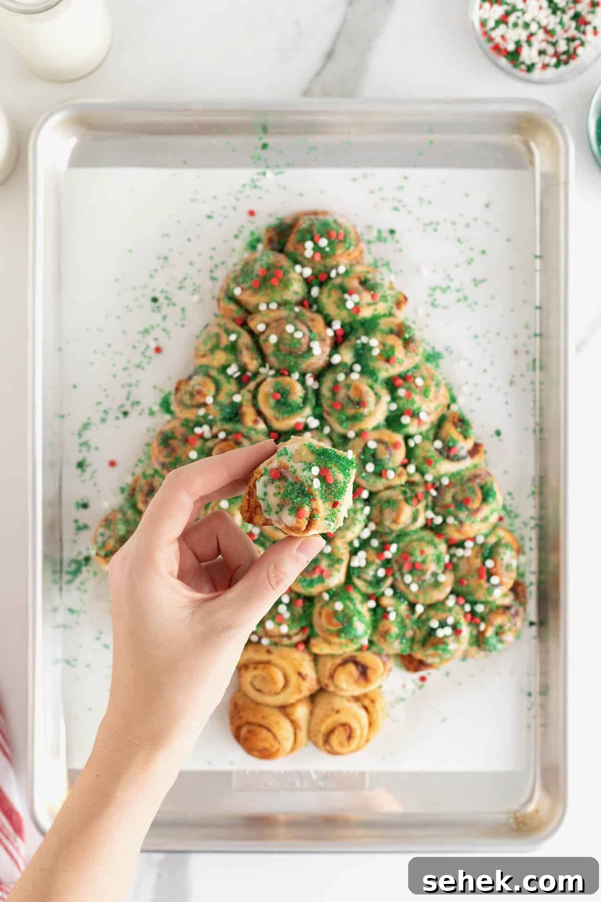 Cinnamon Roll Christmas Tree Pull-Apart Recipe 5 A hand taking a piece of a cinnamon roll pull-apart shaped like a Christmas tree topped with holiday sprinkles.