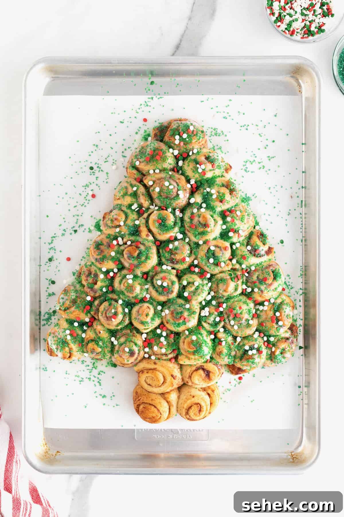Cinnamon Roll Christmas Tree Pull-Apart Recipe 10 A cinnamon roll Christmas tree sprinkled with green sugar crystals and red and green holiday sprinkles on a parchment lined baking sheet.