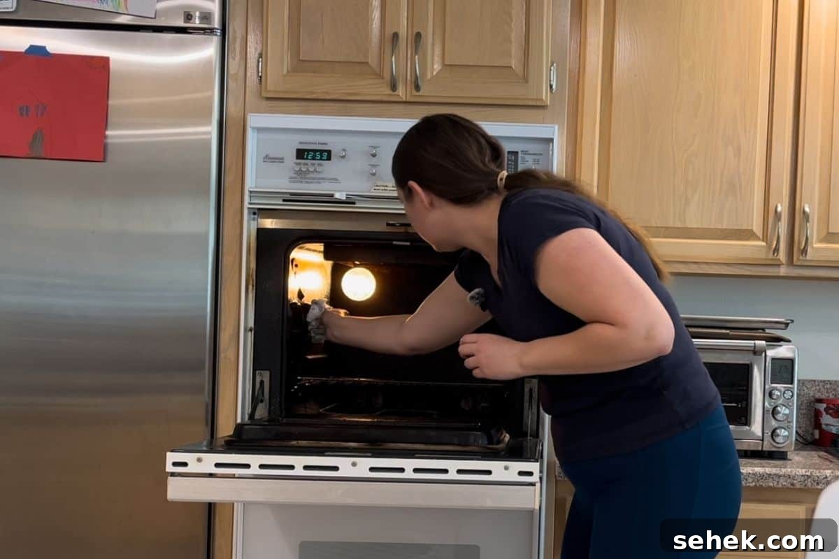 Steam-Clean Your Oven in 2025 Using Simple Kitchen Tools 3 A woman wearing a navy shirt and blue pants is wiping out her oven in a kitchen with wooden cabinets.