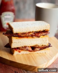 2 bacon sandwich halves stacked on each other with ketchup and cup of tea blurred in background