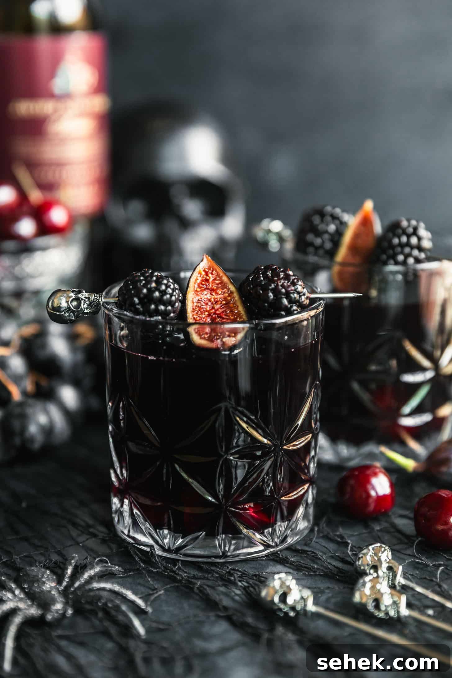 Black Halloween Sangria Recipe with Spooky Blackberry Twist 2 Two glasses of black Halloween sangria garnished with skull picks, blackberries, and figs on a black background next to cherries, grapes. a black skull, and a crystal glass of cherries.