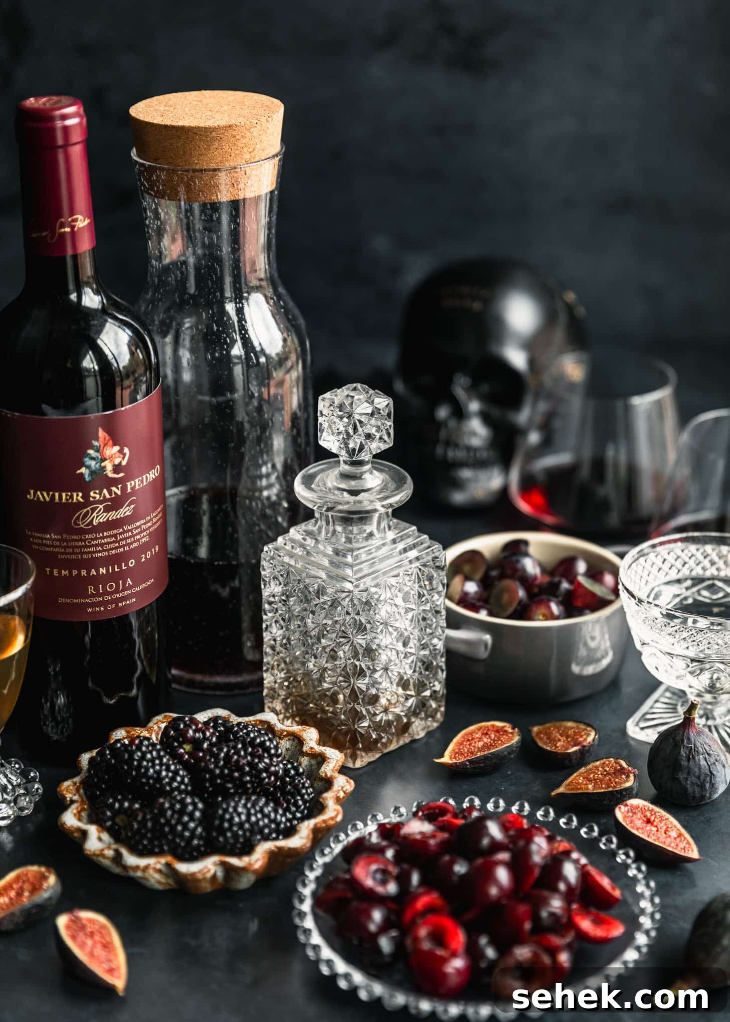 Black Halloween Sangria Recipe with Spooky Blackberry Twist 3 A decanter of brandy, brown bowl of blackberries, pitcher of pomegranate juice, bottle of red wine, grey bowl of grapes, glass plate of cherries, glasses of Triple sec and simple syrup, glasses of red wine, figs, and a black skull on a black background.