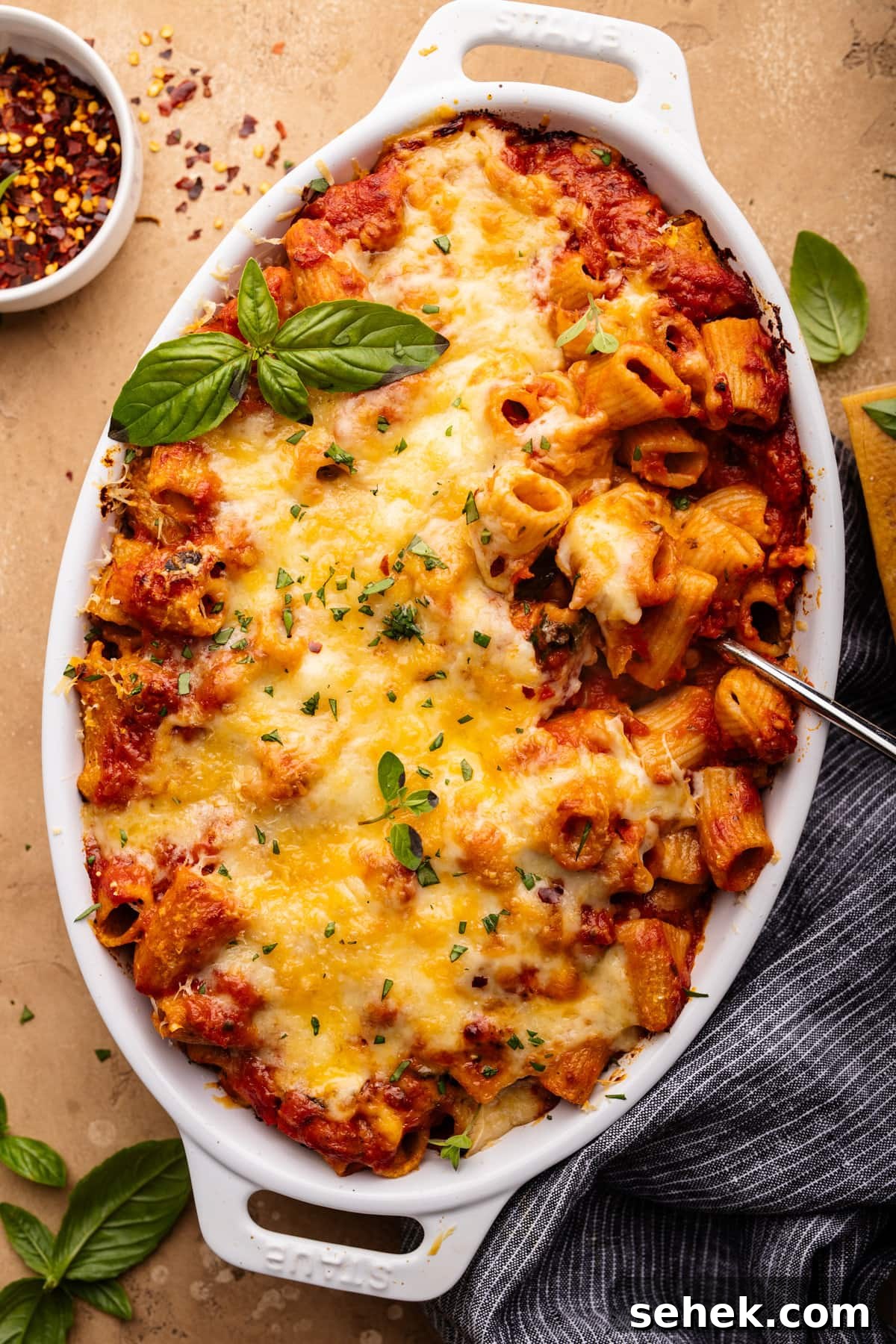 Baked Ziti Without Ricotta: Creamy Oven-Baked Pasta Recipe 2 A white oval-shaped dish with baked ziti and a spoon in it. The dish is sitting on a tan background with fresh herbs, a small bowl of crushed red pepper flakes, and a dark napkin arranged around it.