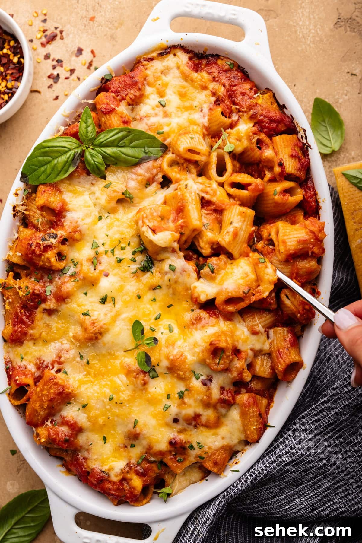 Baked Ziti Without Ricotta: Creamy Oven-Baked Pasta Recipe 8 A white oval-shaped dish with baked ziti in it and a hand lifting a spoon of pasta out of it. The baked ziti is sitting on a tan background with fresh herbs, a small bowl of crushed red pepper flakes, and a dark napkin arranged around it.
