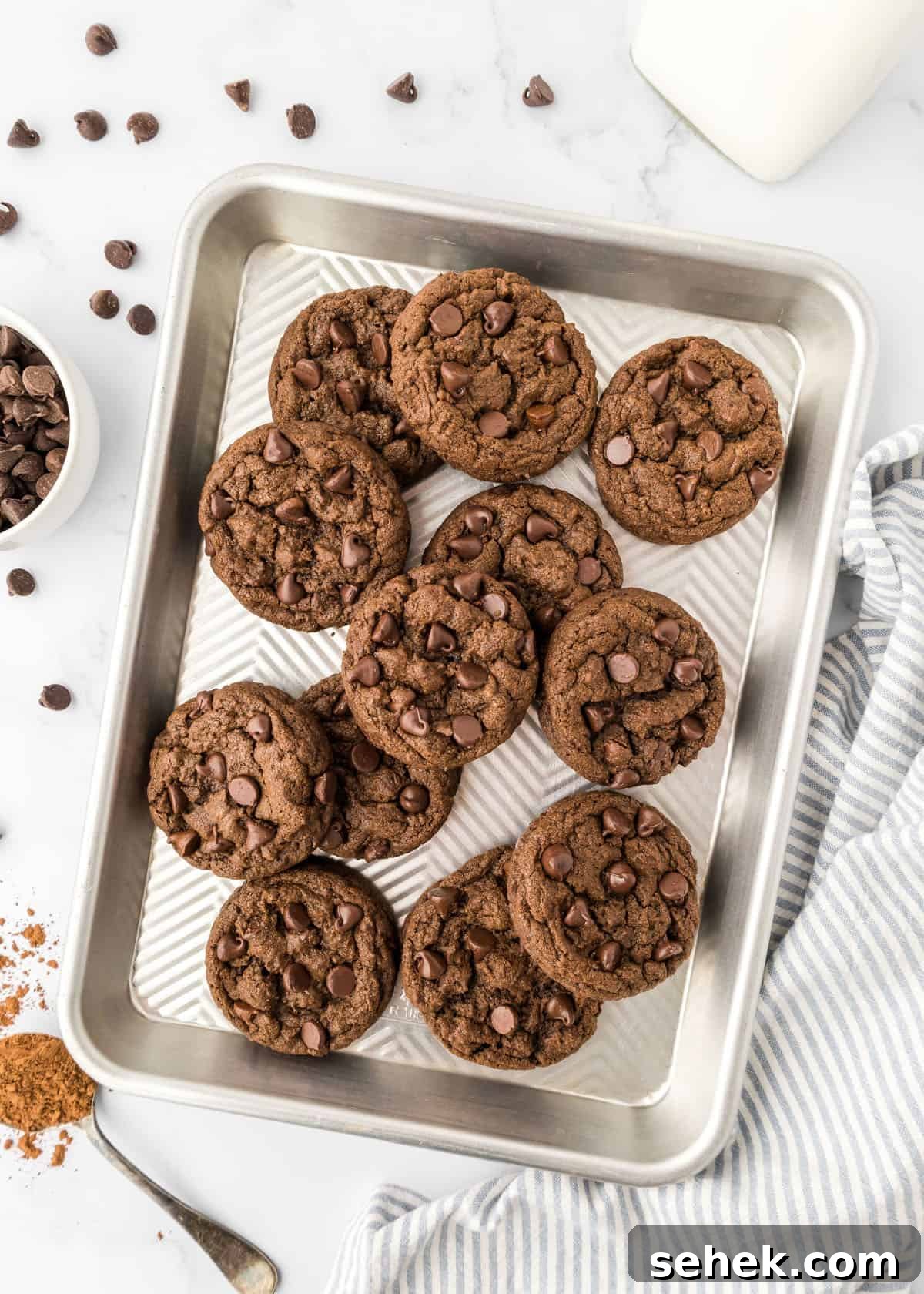 Ultimate Double Chocolate Cookies Recipe for Rich, Fudgy Results 10 Double chocolate chip cookies on a baking sheet on a counter with chocolate chips.
